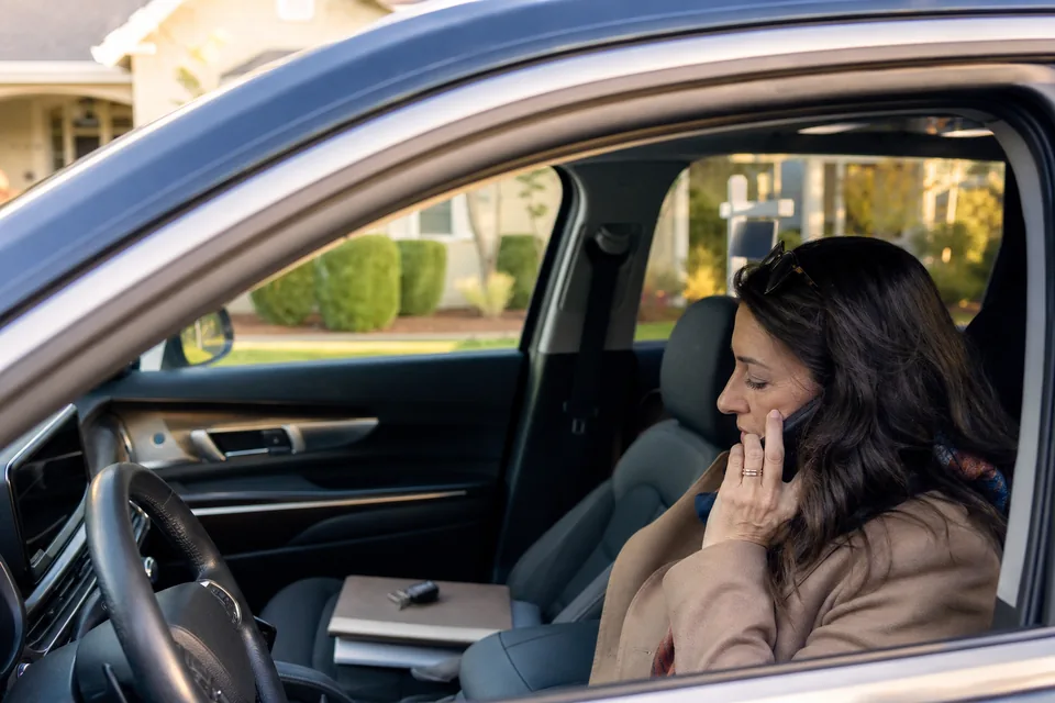 Real estate agent in a parked car answering a transferred lead call on her phone with a listing folder on the passenger seat