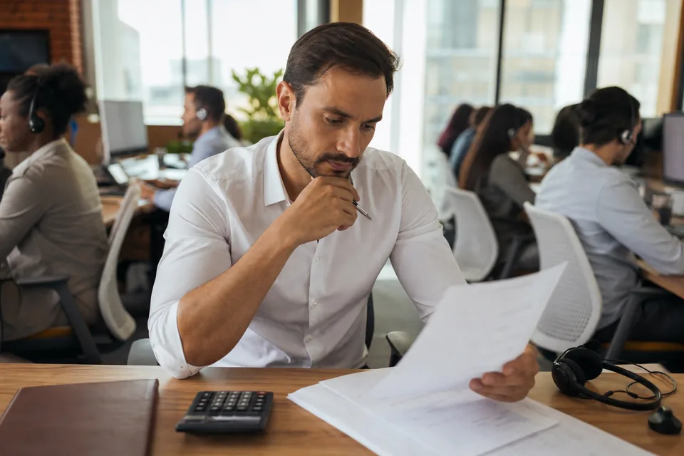 Call center owner reviewing an annual dialer contract with a calculator while agents make outbound calls behind him
